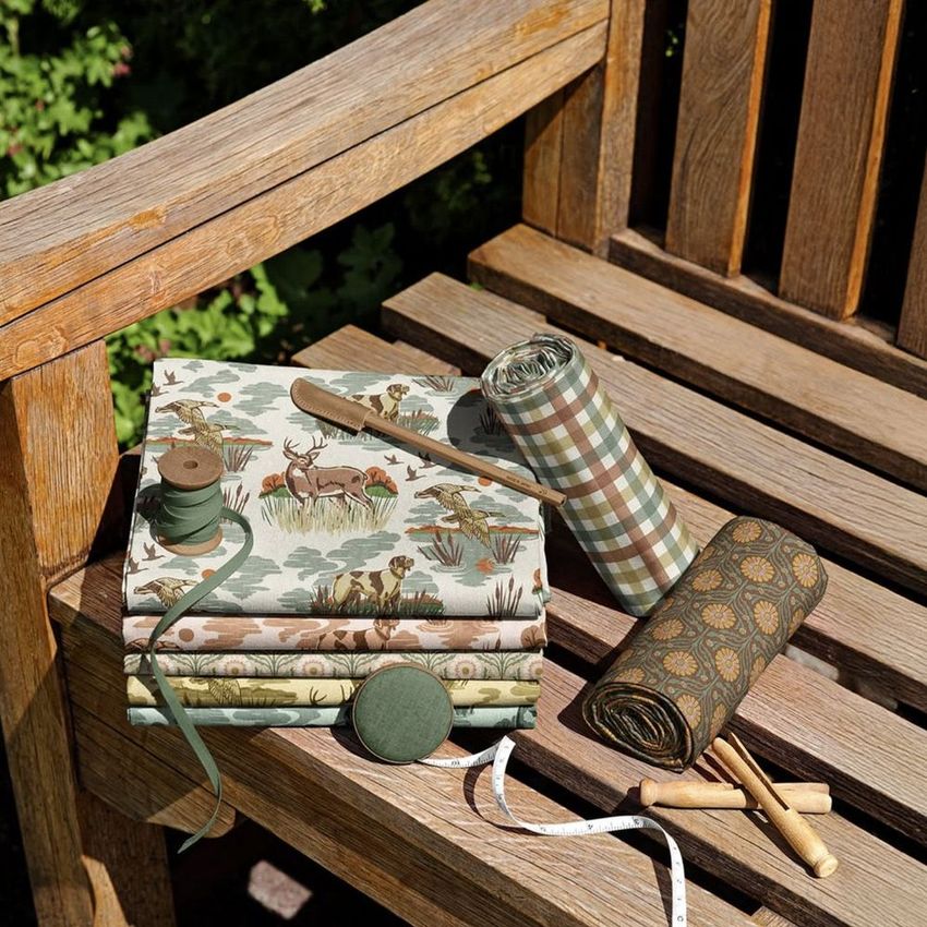 Fabric rolls and folded fabrics with autumn-themed patterns displayed on a wooden garden bench, surrounded by sewing tools.