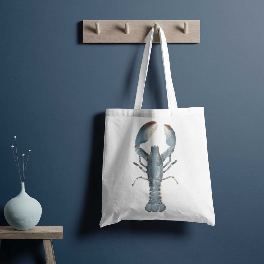 Tote bag mockup featuring a white bag with a watercolor-style blue lobster illustration, hanging on a wooden wall hook.