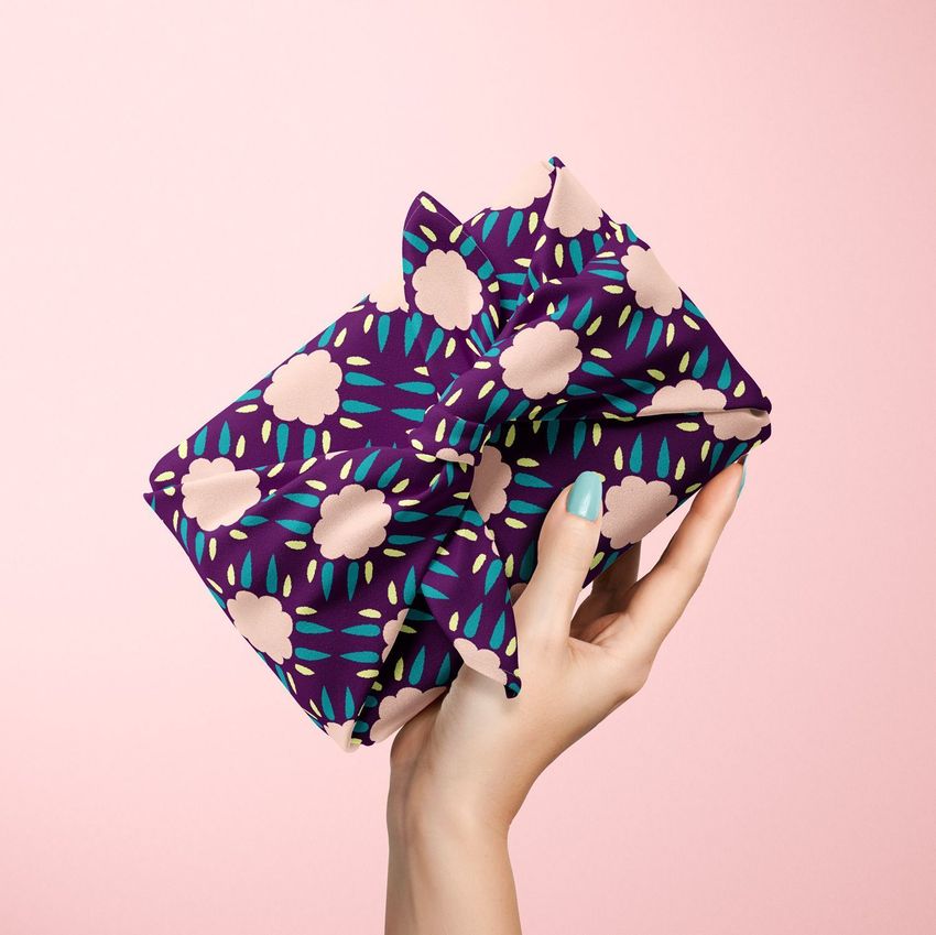 Hand holding a furoshiki fabric wrap mockup with a purple base, floral pattern, and teal accents against a pink background.