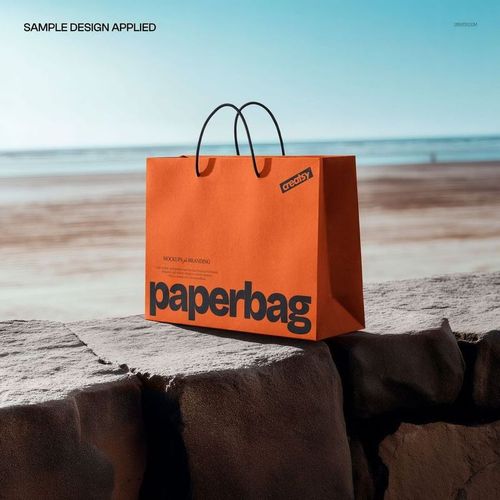 Orange shopping bag mockup with black lettering and handles, shown from the front on a rocky surface by the ocean.