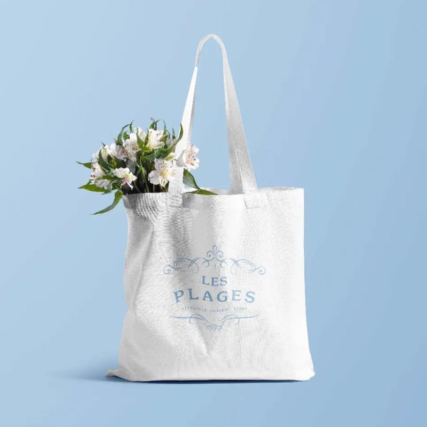 White tote bag mockup with blue decorative text and floral graphic, filled with white flowers, on a light blue background.