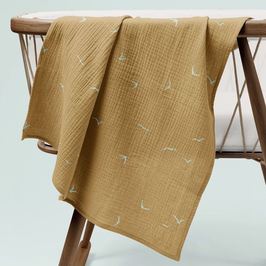 Crinkle muslin swaddle blanket mockup draped over a wooden crib, featuring a mustard yellow fabric with a simple bird pattern.