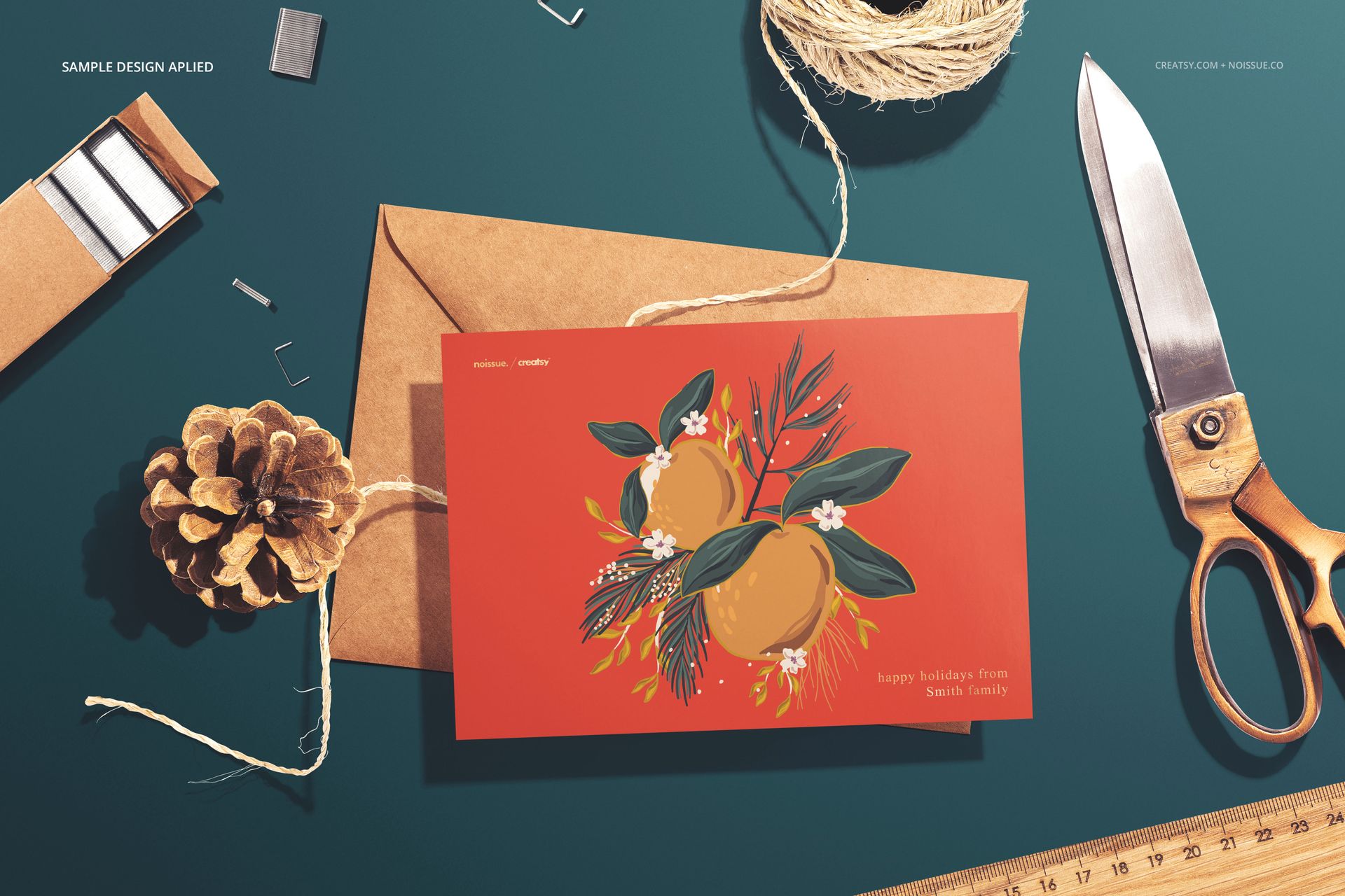 Stationery mockup featuring a red greeting card with orange fruit illustration, brown envelope, scissors, and twine on a teal surface.
