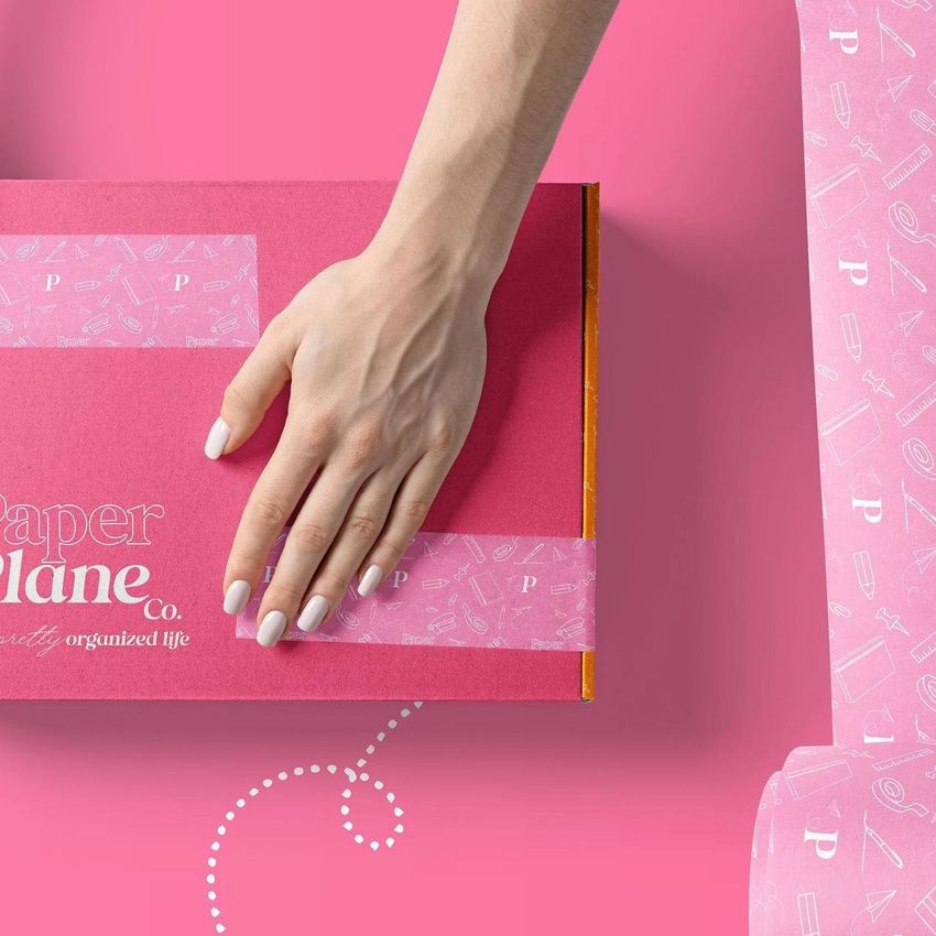 Mailing box mockup featuring a hand pressing patterned tape onto a pink box with white text, set against a pink background.