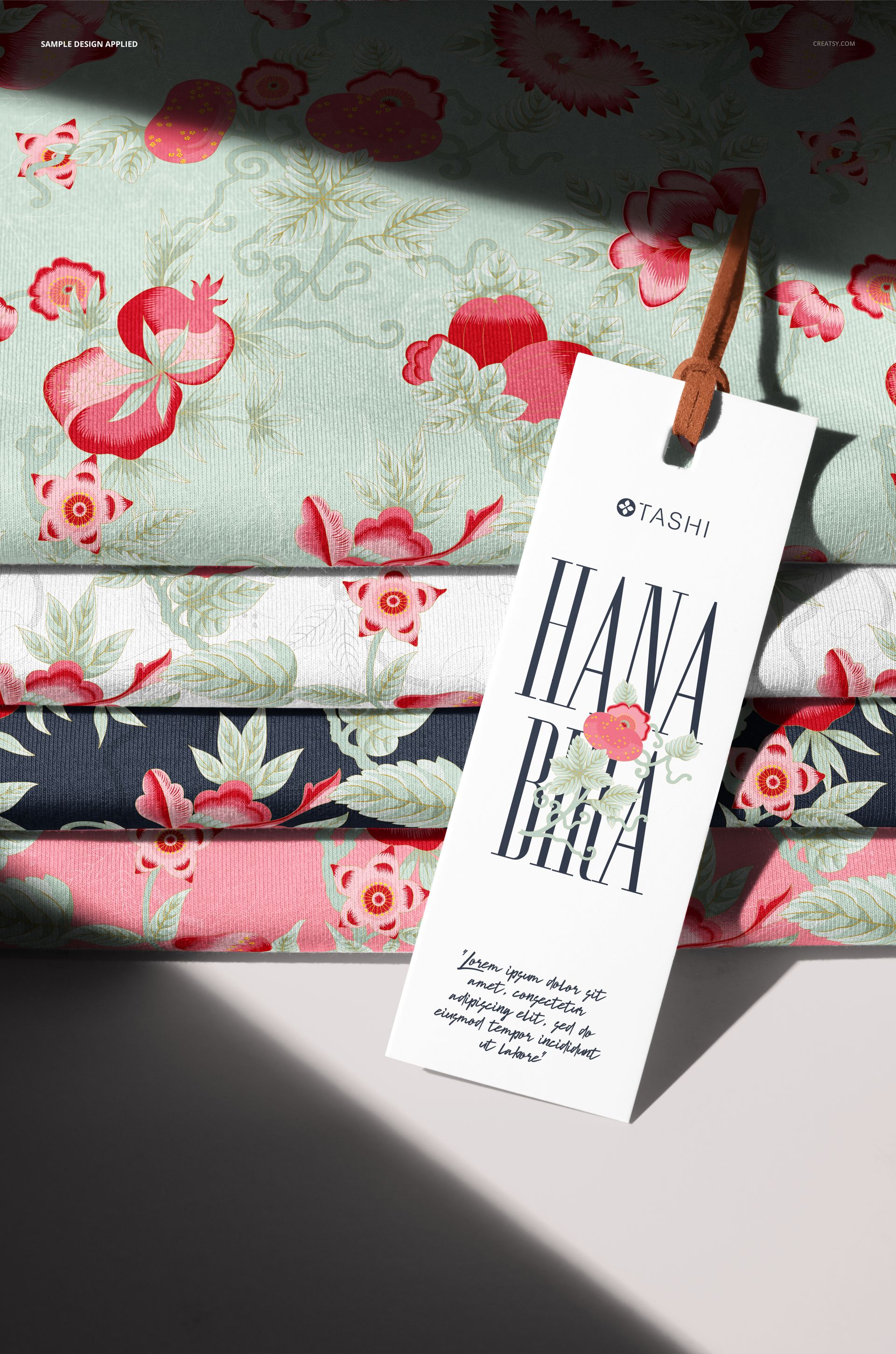 PSD mockup showing a pile of floral jersey fabrics in various colors, with a white branding tag attached by a string.