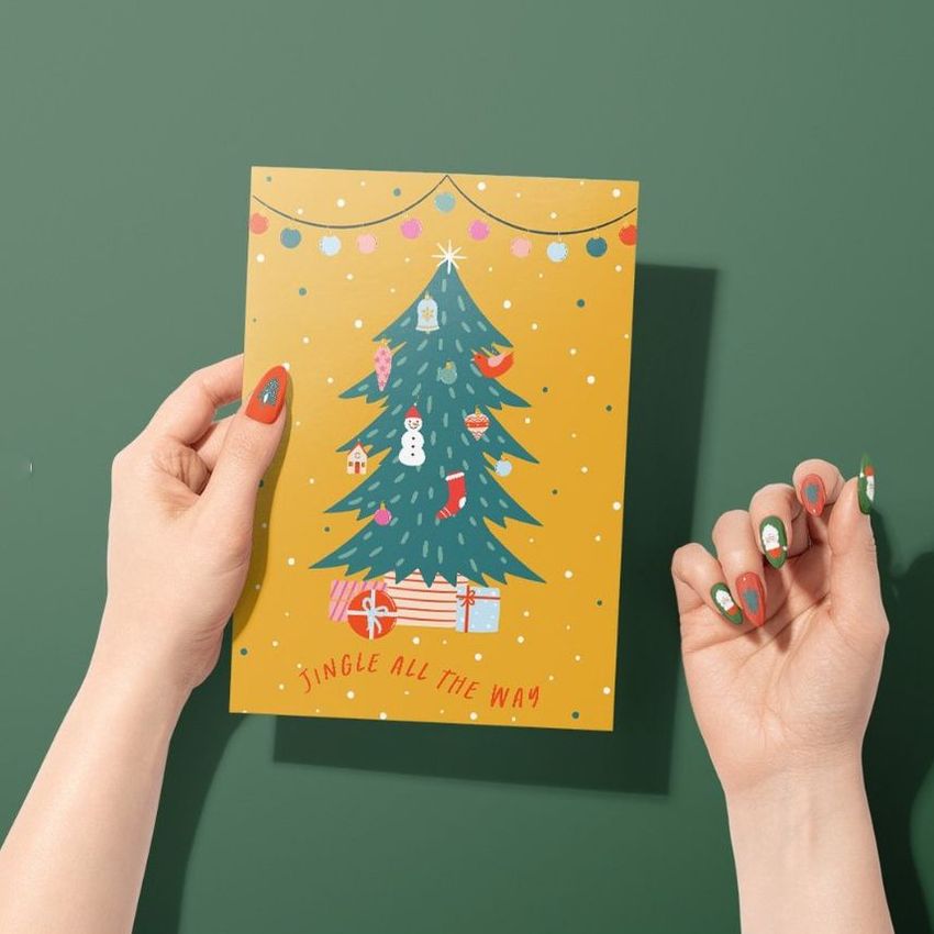 Greeting card mockup featuring a woman's hands holding a yellow card with a decorated Christmas tree and gifts on a green background.