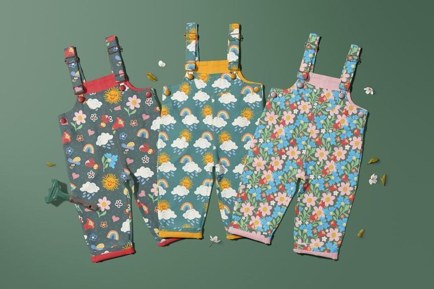 Kids dungarees mockup featuring three pairs with colorful nature-inspired patterns on a green background with leaves and props.