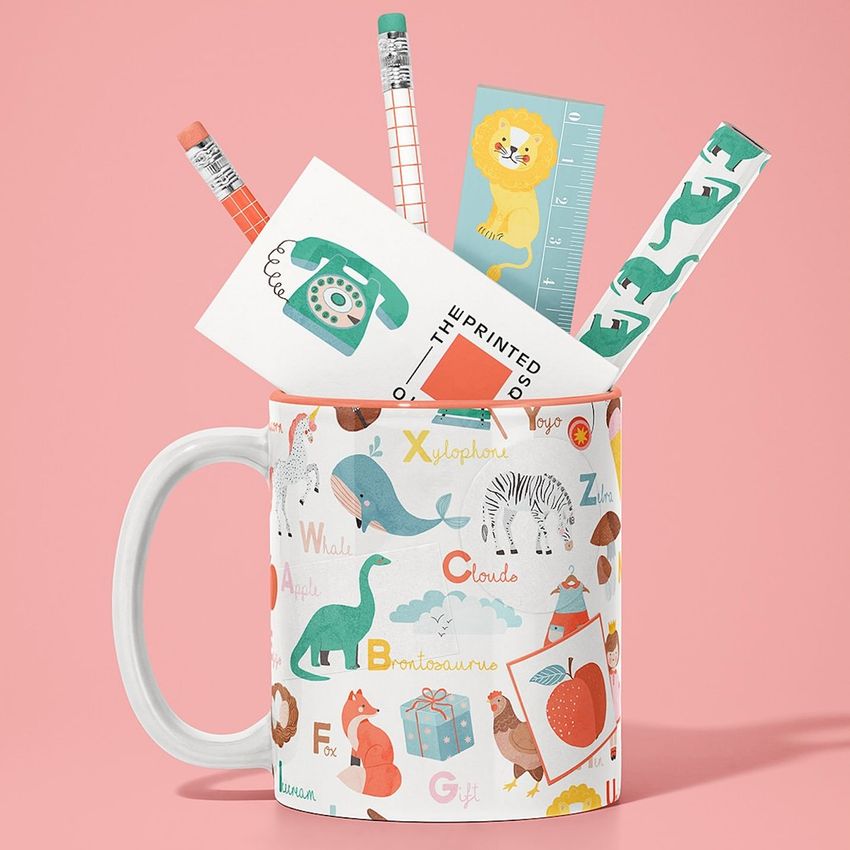 Mug mockup with colorful alphabet animal illustrations, holding pencils, a ruler, and stationery items on a pink background.