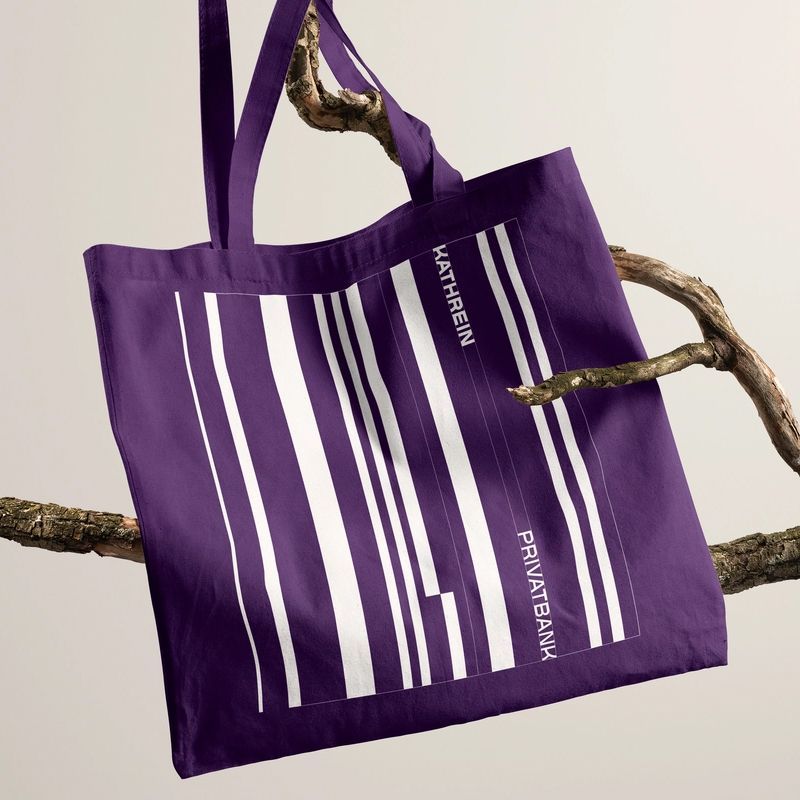 Purple tote bag mockup displayed hanging on a branch, featuring bold white vertical lines and readable text on the front.