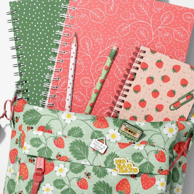 School backpack mockup with a strawberry and daisy pattern, surrounded by spiral notebooks and stationery in red and green tones.