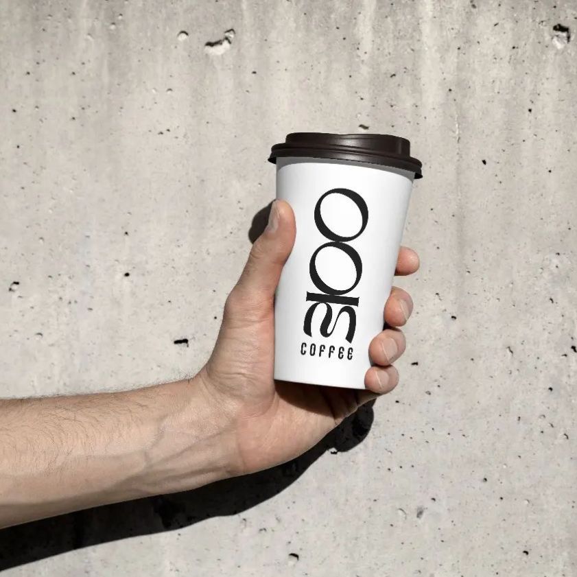 Hand holding a white paper cup mockup with bold black vertical text, set against a textured concrete wall in natural light.