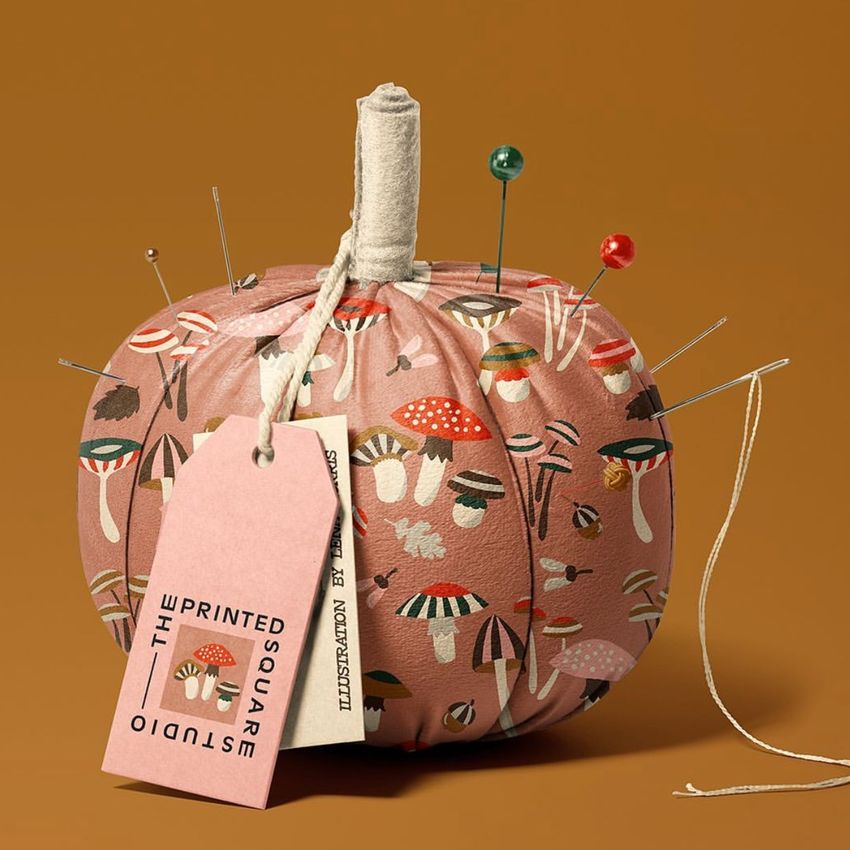 Fabric pumpkin mockup with a pink mushroom pattern, used as a pincushion with several pins and a tag attached, on a brown background.