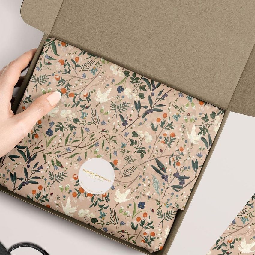Mailer box mockup featuring floral pattern tissue paper in soft earthy tones, partially unwrapped by a hand on a neutral background.