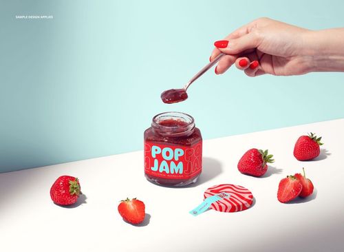 PSD mockup featuring a jam jar with bold blue and red label, strawberries, and a hand lifting jam with a spoon.