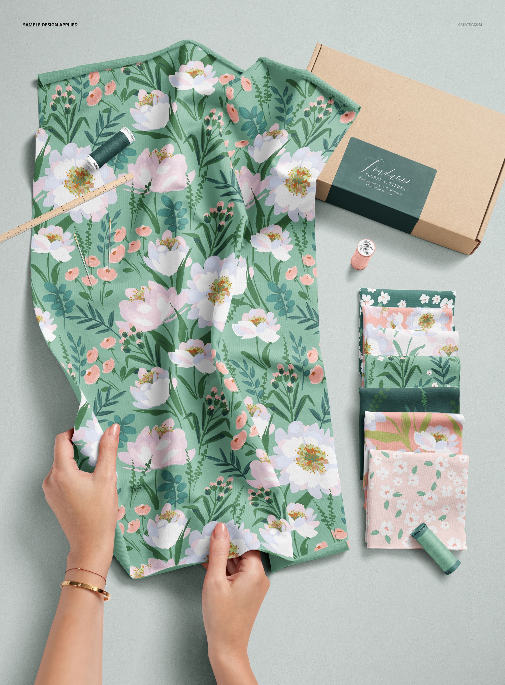 Top view of green floral jersey fabric with sewing supplies, hands arranging cloth, and a cardboard box mockup.