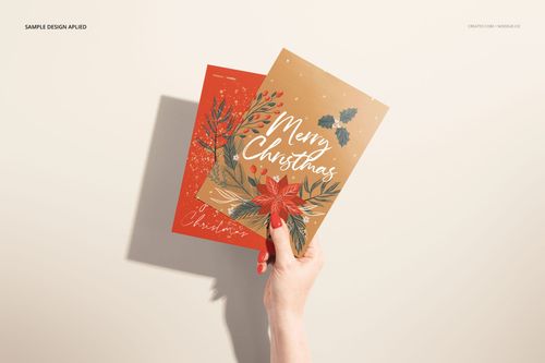 Person’s hand displaying two mockup cards featuring Christmas-themed illustrations and text, top view on beige surface.