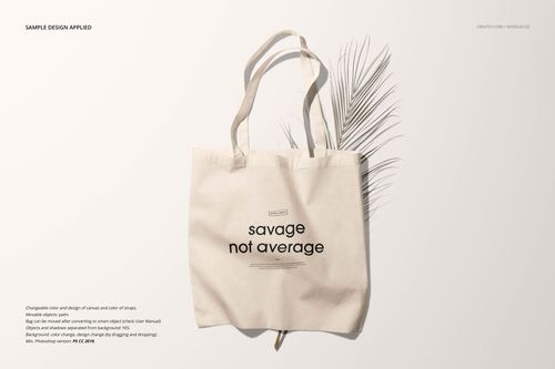 Front view of a beige tote bag mockup with black text design, placed on a light surface with a dried palm leaf.