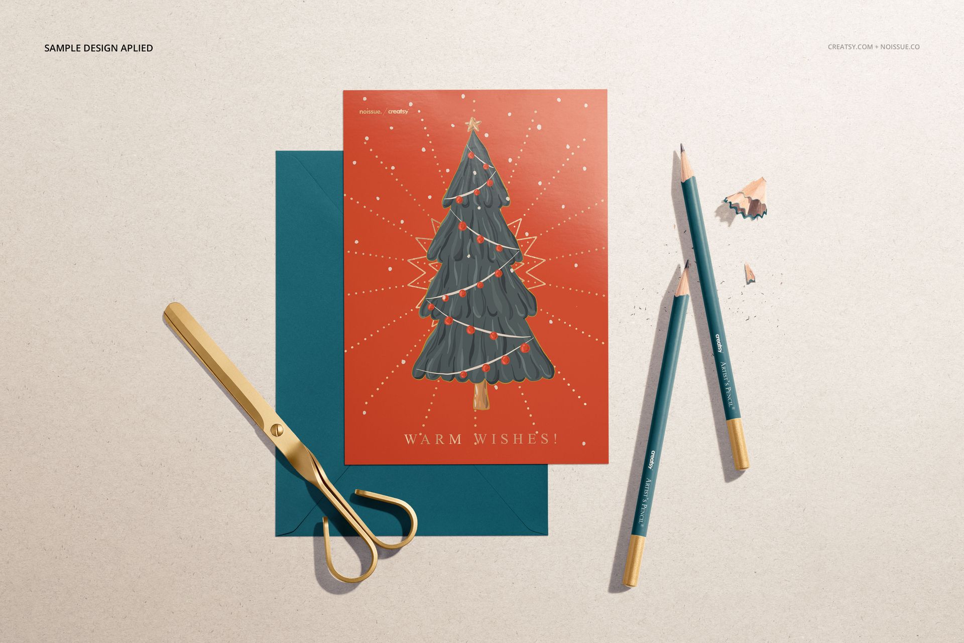 Stationery mockup featuring a holiday card with a tree illustration, teal envelope, pencils, and gold scissors on a beige surface.