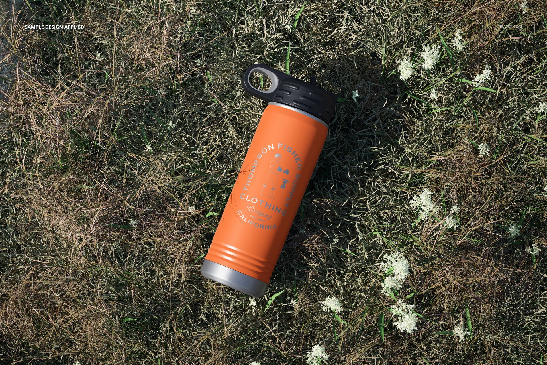 Powder coated orange water bottle mockup with black cap placed on grassy ground, surrounded by small white flowers.