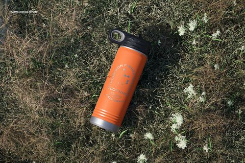 Powder coated orange water bottle mockup with black cap placed on grassy ground, surrounded by small white flowers.