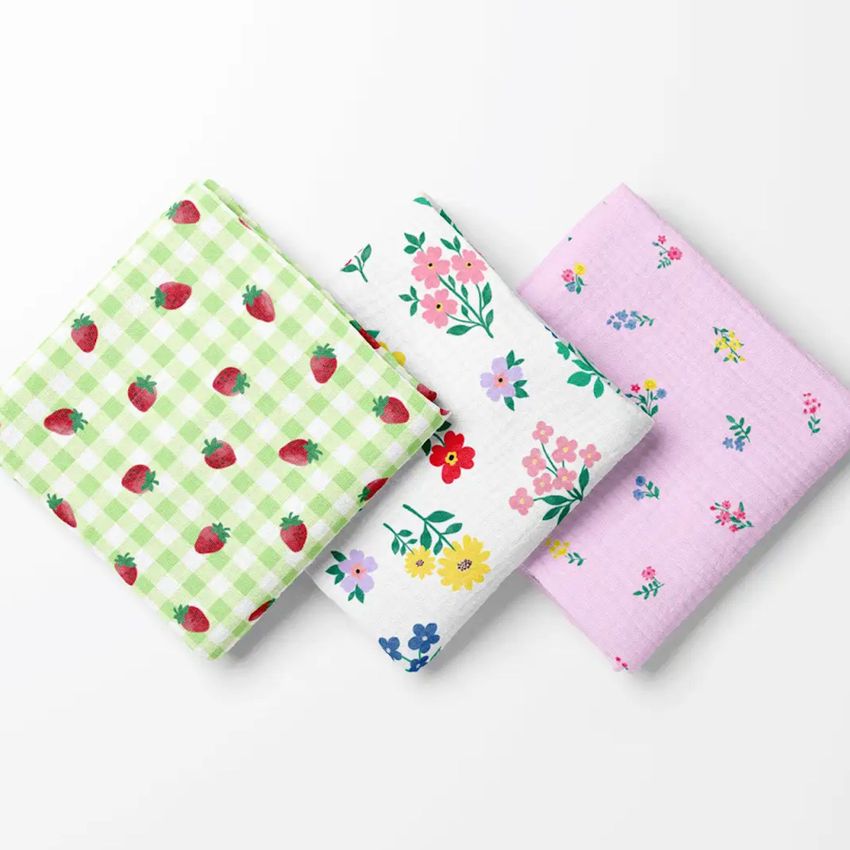 Three crinkle muslin swaddle blankets are folded side by side, each featuring a different colorful floral or fruit pattern.