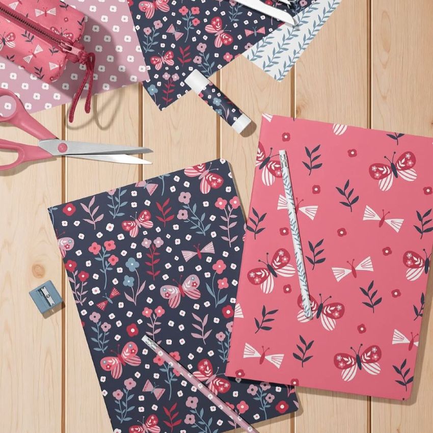 Stationery mockup featuring two A4 notebooks with butterfly and floral patterns, pencils, scissors, and a sharpener on a wooden surface.