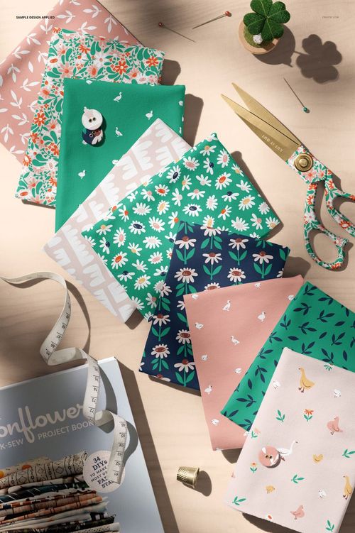 Folded jersey fabrics in various pastel and green tones with floral and animal patterns, shown from above on a table with sewing supplies.