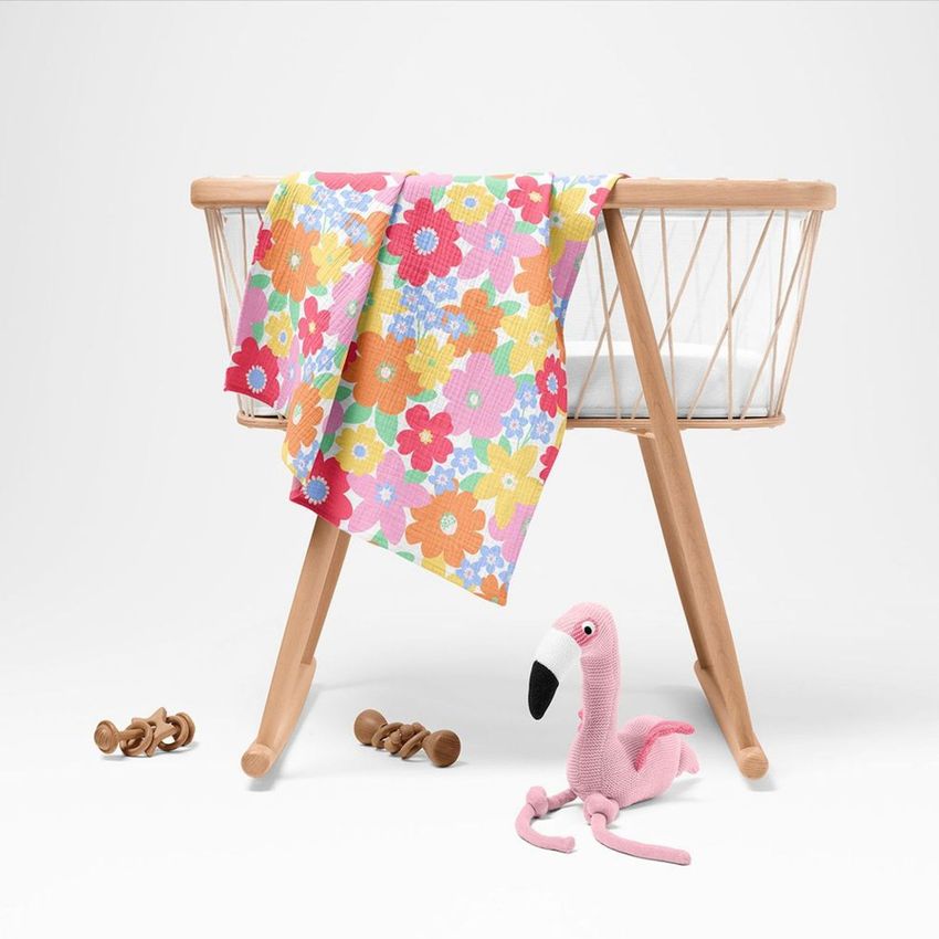 Crinkle muslin swaddle blanket mockup draped over a light wooden bassinet, featuring a colorful floral pattern.