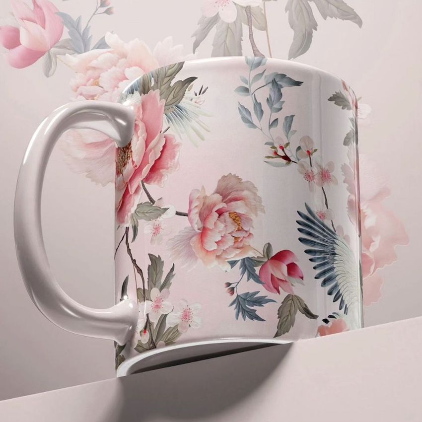 Mug mockup featuring a close-up of a white ceramic mug with a pink floral and bird pattern, set on a white block with a matching floral background.