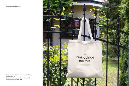 Neutral canvas tote bag mockup hanging on a black wrought iron fence outdoors, surrounded by green foliage.