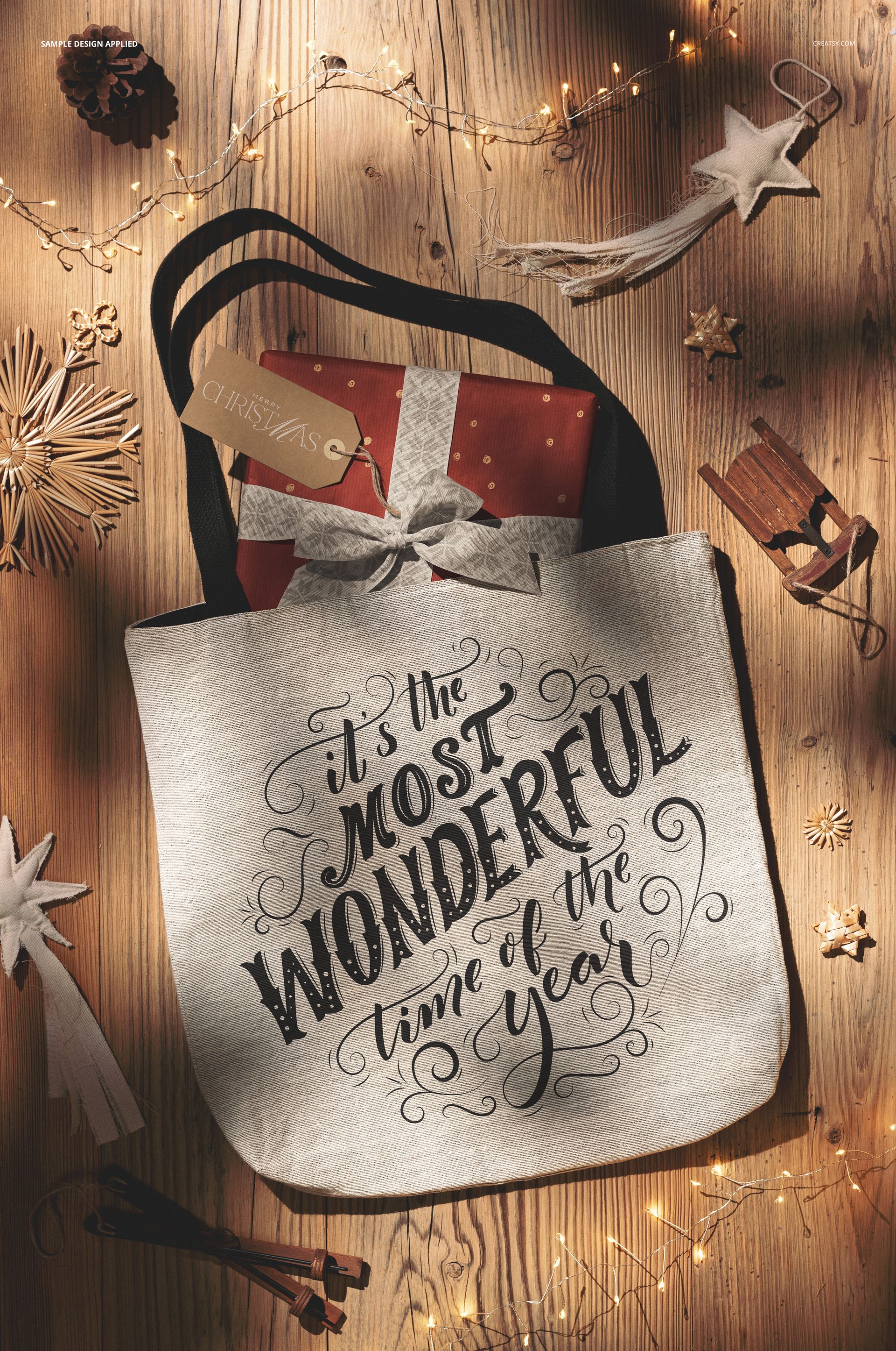 Top view of a Christmas-themed tote bag mockup with black handles, holiday lights, and seasonal props on wood.