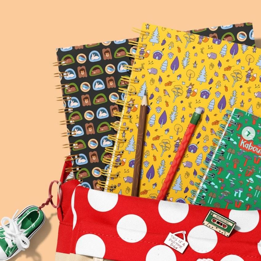 School backpack mockup with spiral notebooks featuring playful woodland patterns, a pencil, and a red polka dot pouch on a peach background.