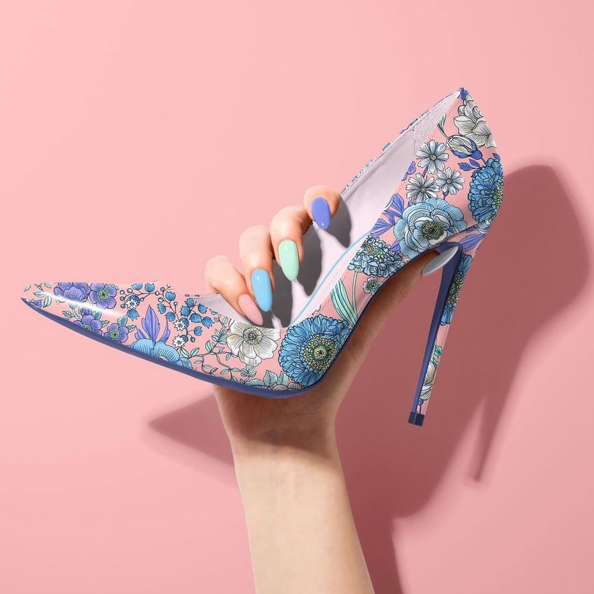 High heels mockup featuring a hand holding a pointed shoe with a blue and pink floral pattern against a pastel pink background.