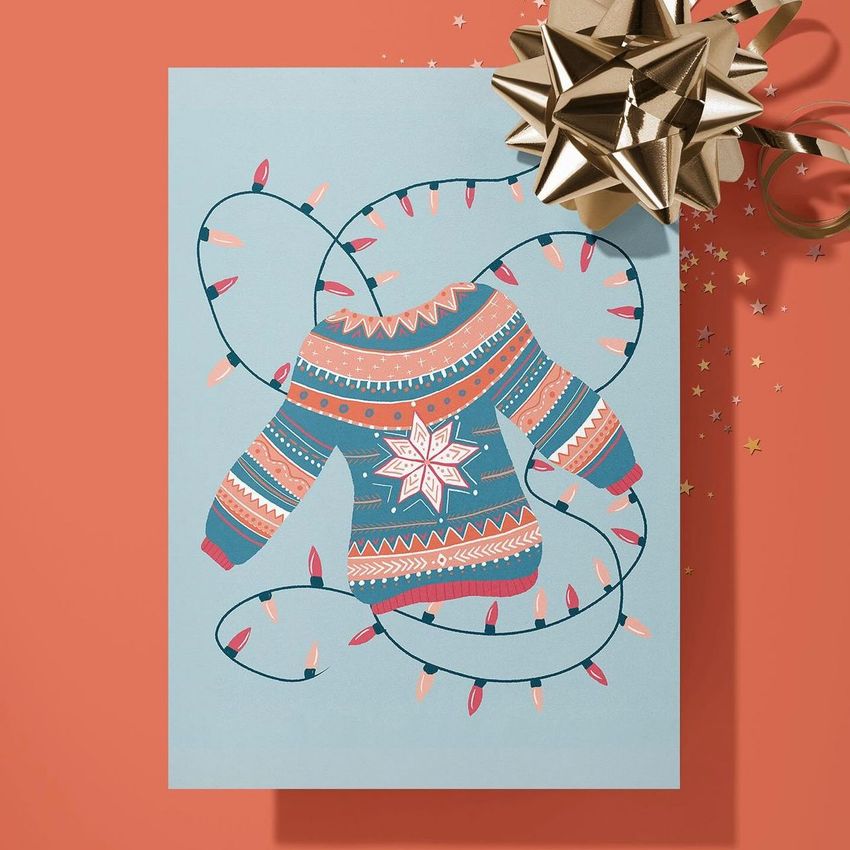 Greeting card mockup featuring an illustrated sweater with festive patterns and holiday lights on a blue background, set on a peach surface.