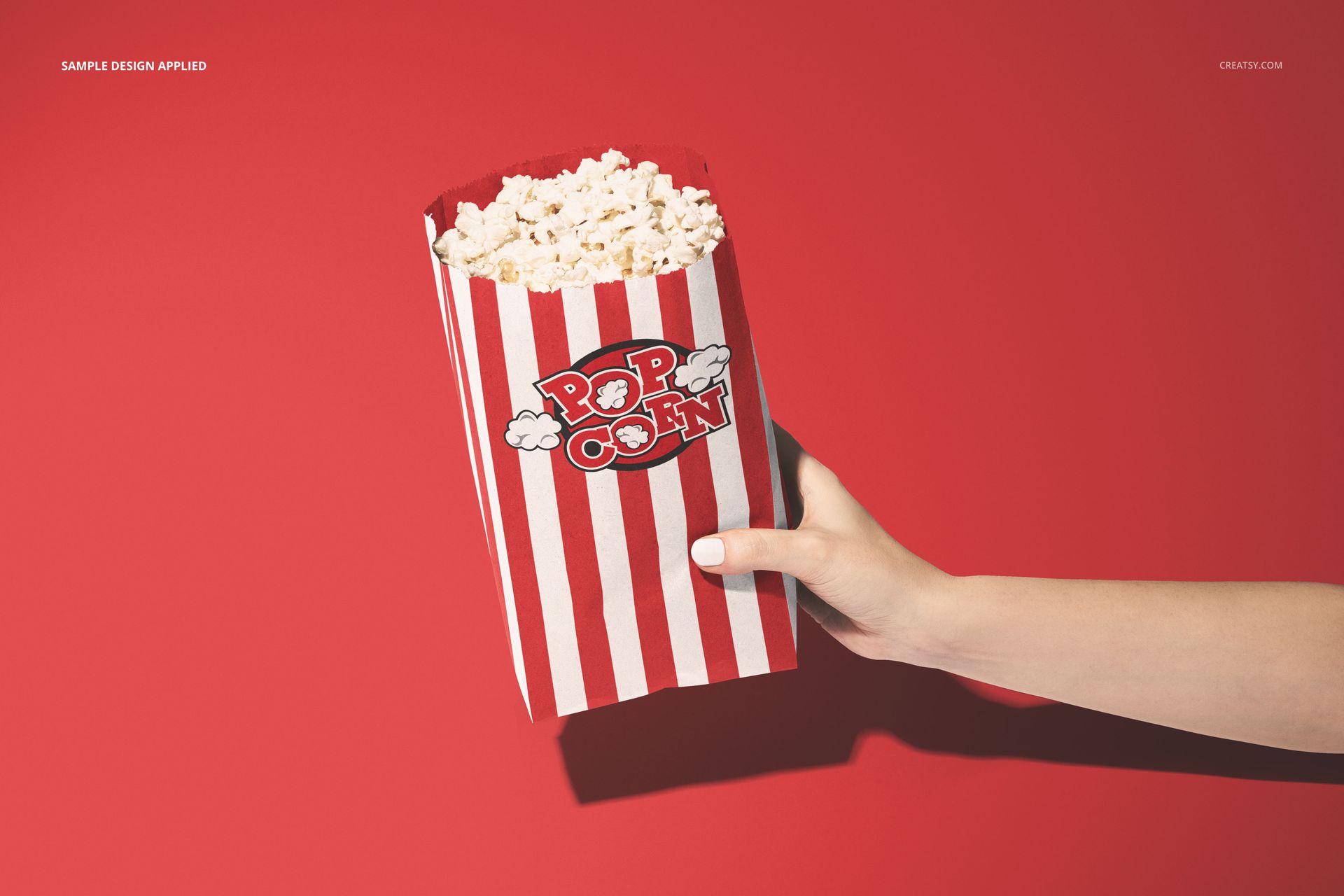 Front view of a popcorn paper bag mockup with vertical red and white stripes, held by a hand on a red backdrop.