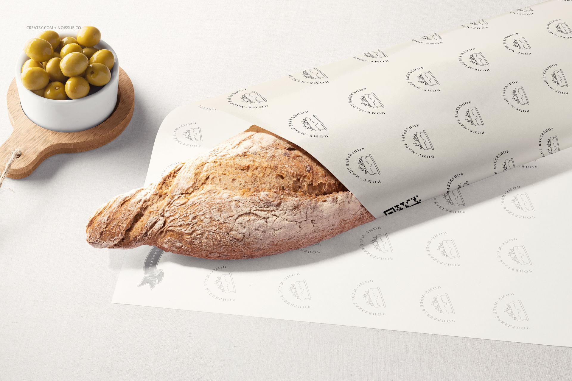 PSD mockup scene featuring a loaf of bread in printed food safe paper, accompanied by a small bowl of green olives.