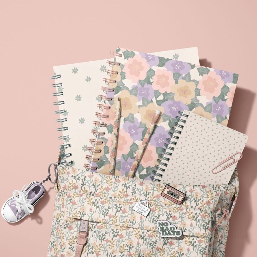 School backpack mockup with spiral notebooks, pastel floral covers, and stationery items arranged on a soft pink background.