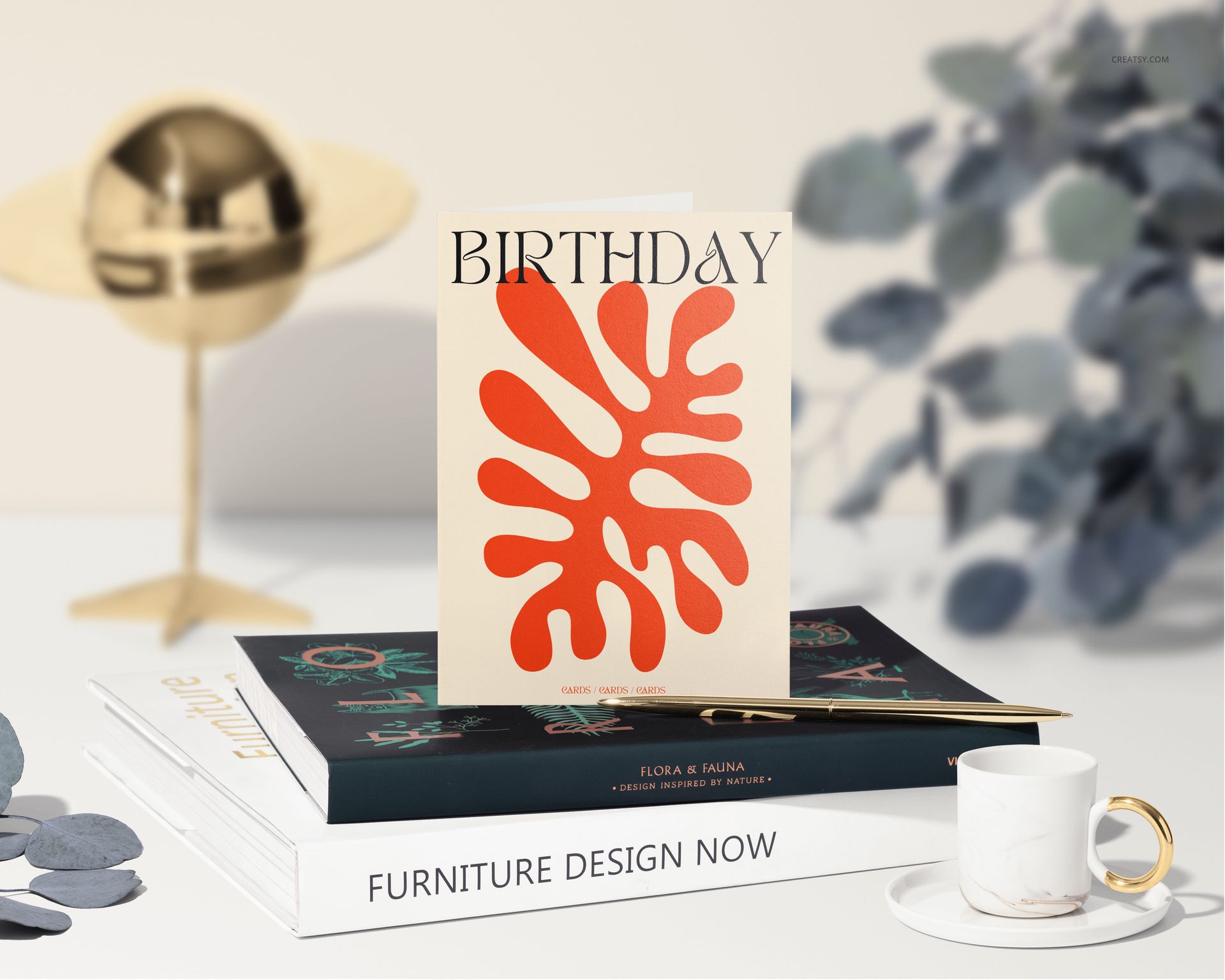 Greeting card mockup featuring a cream card with orange abstract pattern and gold pen, set on decorative books.