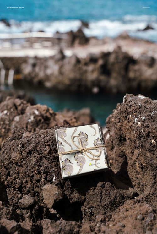 PSD mockup showing patterned fabric wrapped with twine, displayed outdoors on rocky shoreline by blue water.
