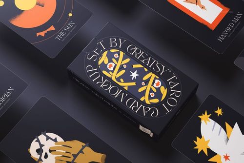 Tarot cards and a rectangular box displayed on a dark surface, featuring bold graphic illustrations in orange and yellow tones.