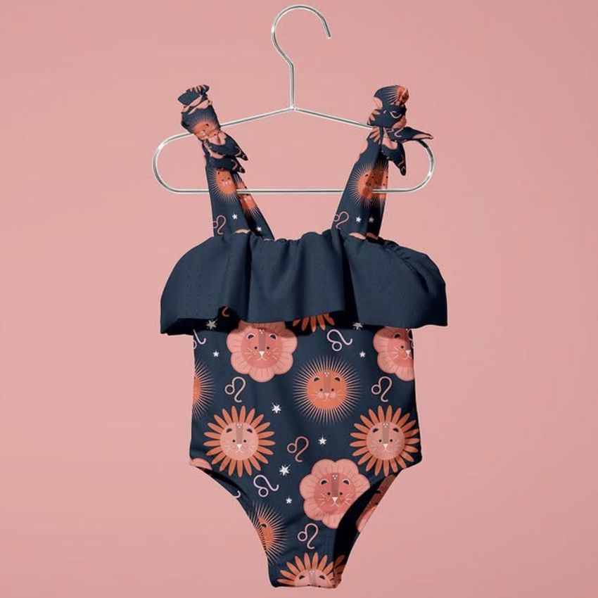 Baby swimsuit mockup featuring a ruffled navy blue one-piece with playful orange sun and lion illustrations, displayed on a hanger against a pink background.