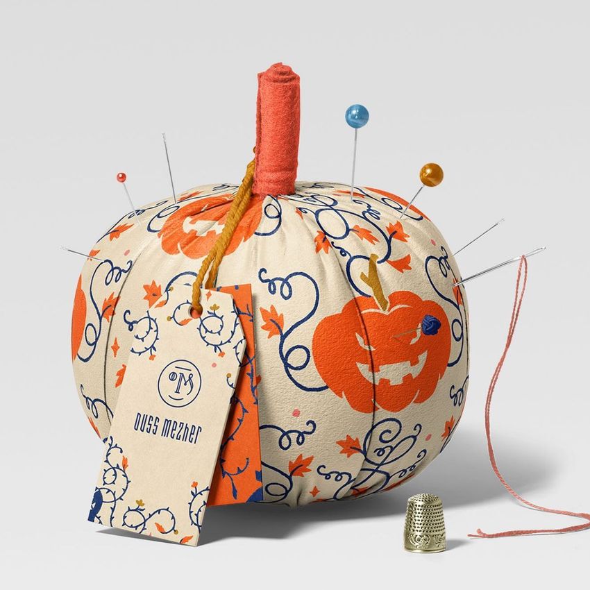 Fabric pumpkin mockup with an orange and blue Halloween pattern, used as a pincushion with sewing pins and a tag attached.