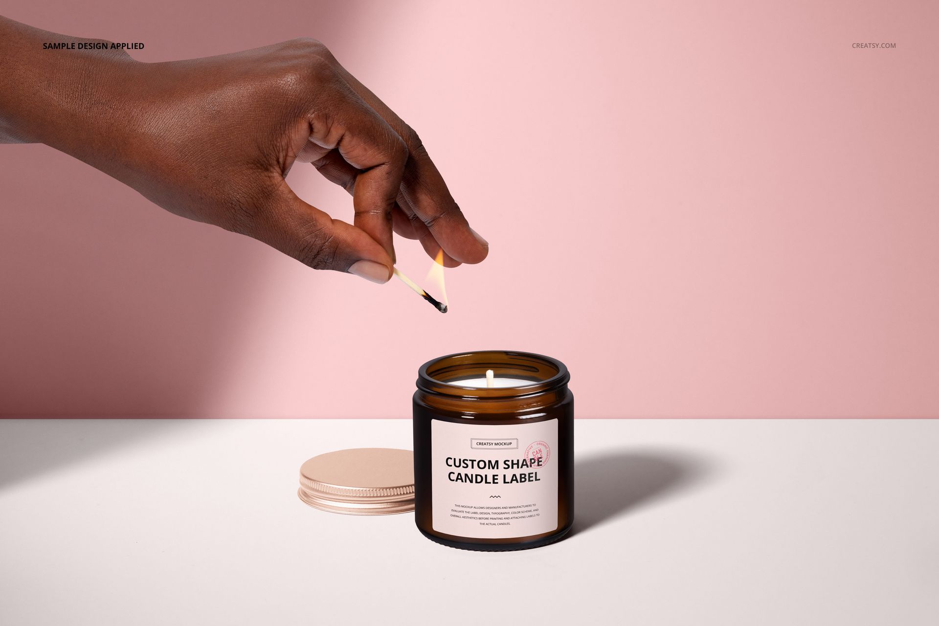 Front view of an amber jar candle mockup with cotton wick, hand holding match above, lid placed nearby, pink wall.
