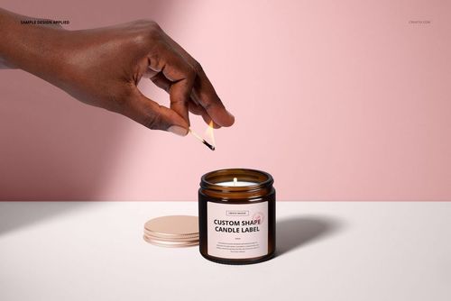 Front view of an amber jar candle mockup with cotton wick, hand holding match above, lid placed nearby, pink wall.