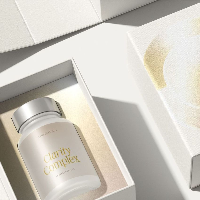 Cosmetics box packaging mockup featuring a white capsule bottle labeled 'Clarity Complex' in an open box on a light surface.