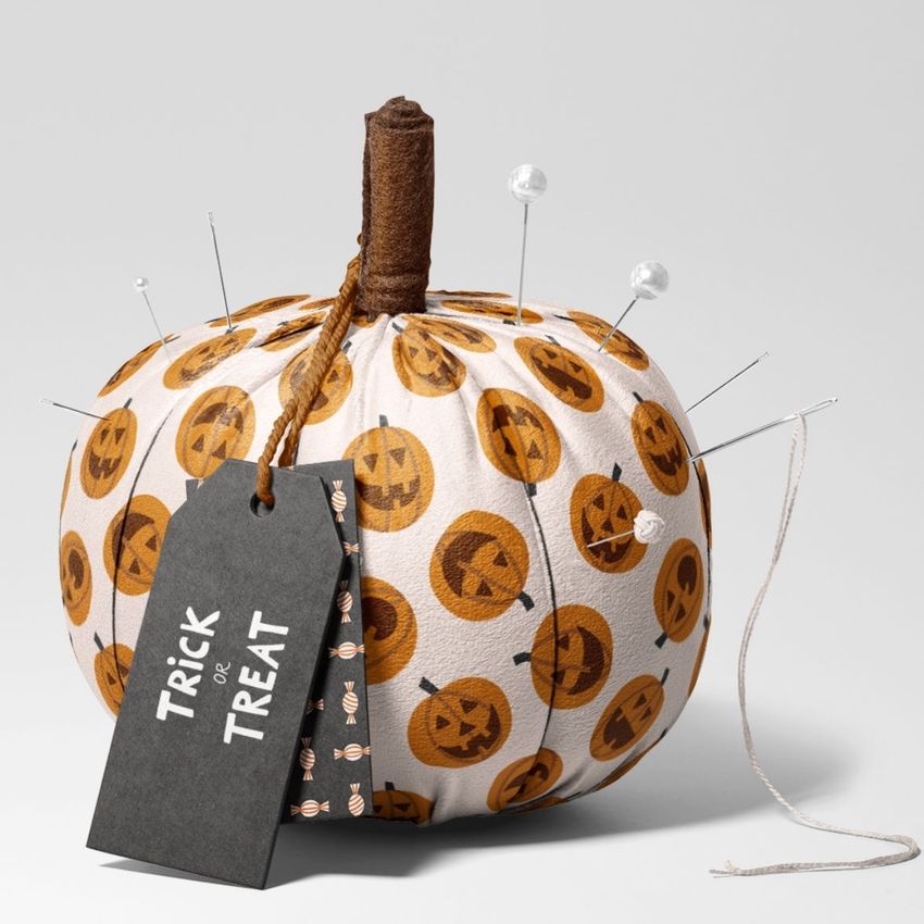 Fabric pumpkin mockup featuring a white textile pumpkin with orange jack-o'-lantern patterns, pins, and a black tag reading 'Trick or Treat'.