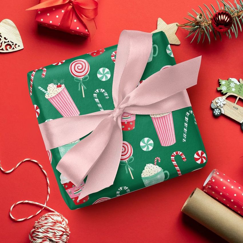 Square gift box mockup wrapped in festive paper with candy cane and peppermint patterns, tied with a pink ribbon on a red background.