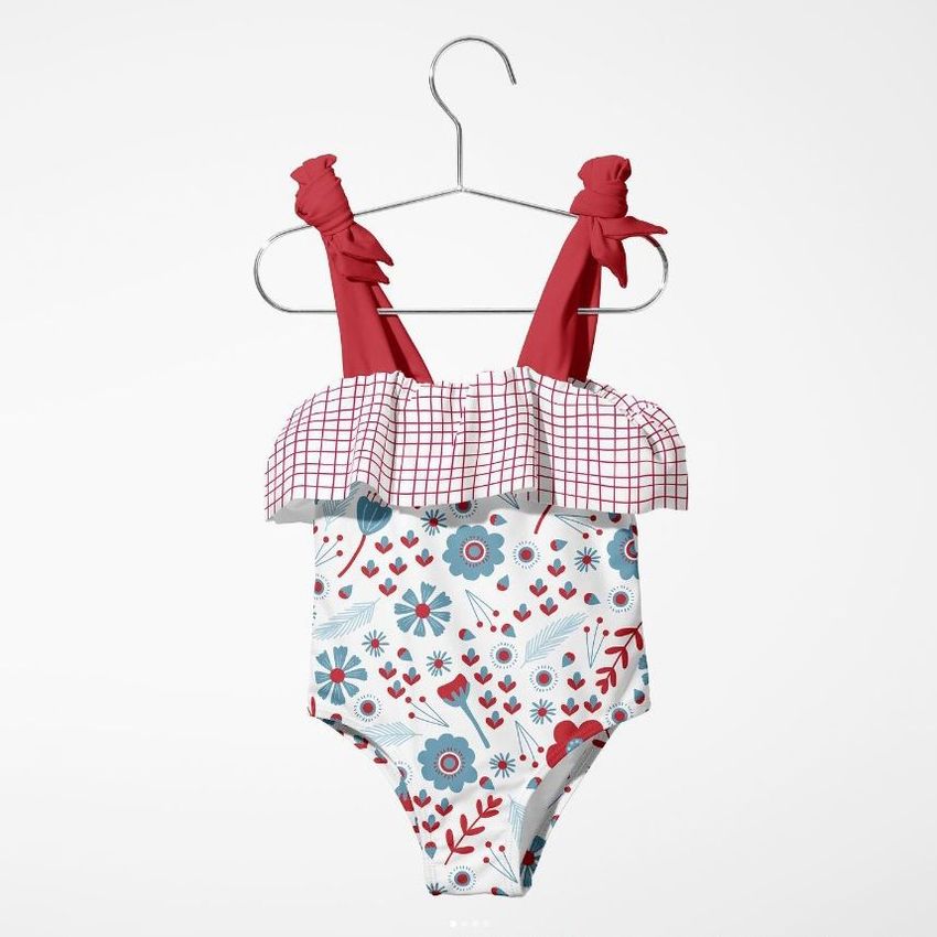 Baby swimsuit mockup featuring a floral pattern with blue and red flowers, red shoulder straps, and a checkered ruffle, displayed on a hanger against a plain white background.