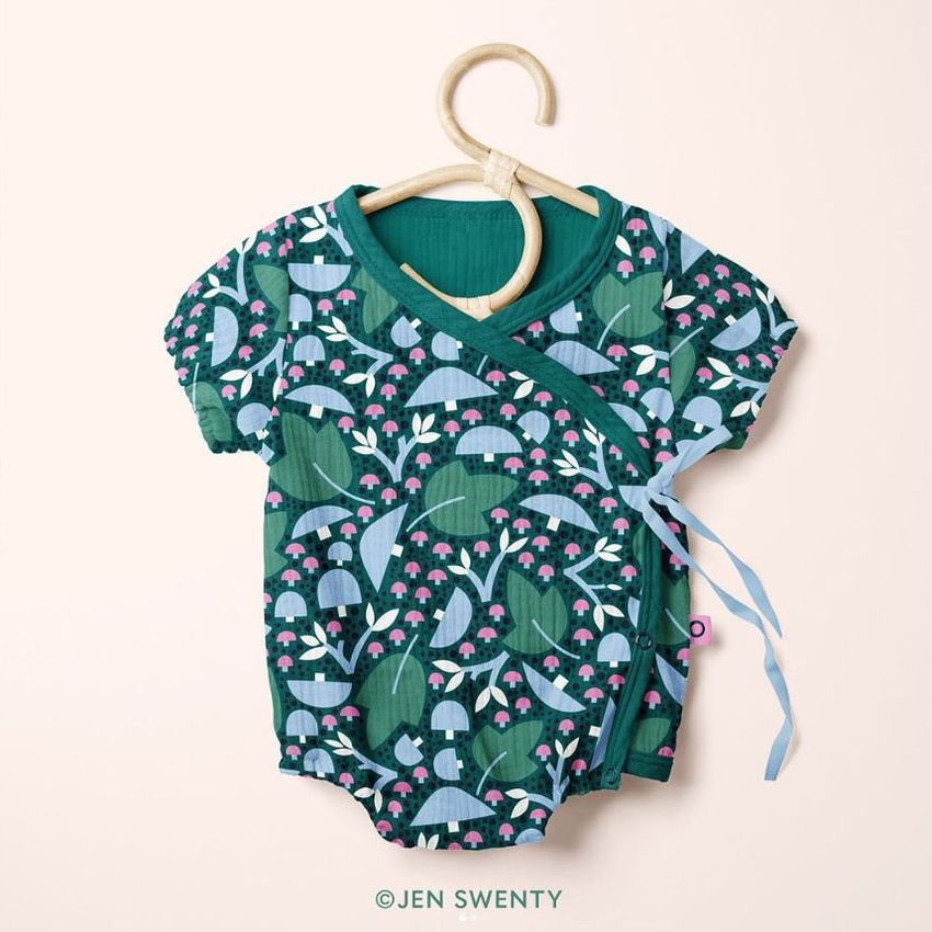 Muslin onesie mockup featuring a green baby bodysuit with a whimsical mushroom and foliage pattern, displayed on a wooden hanger.
