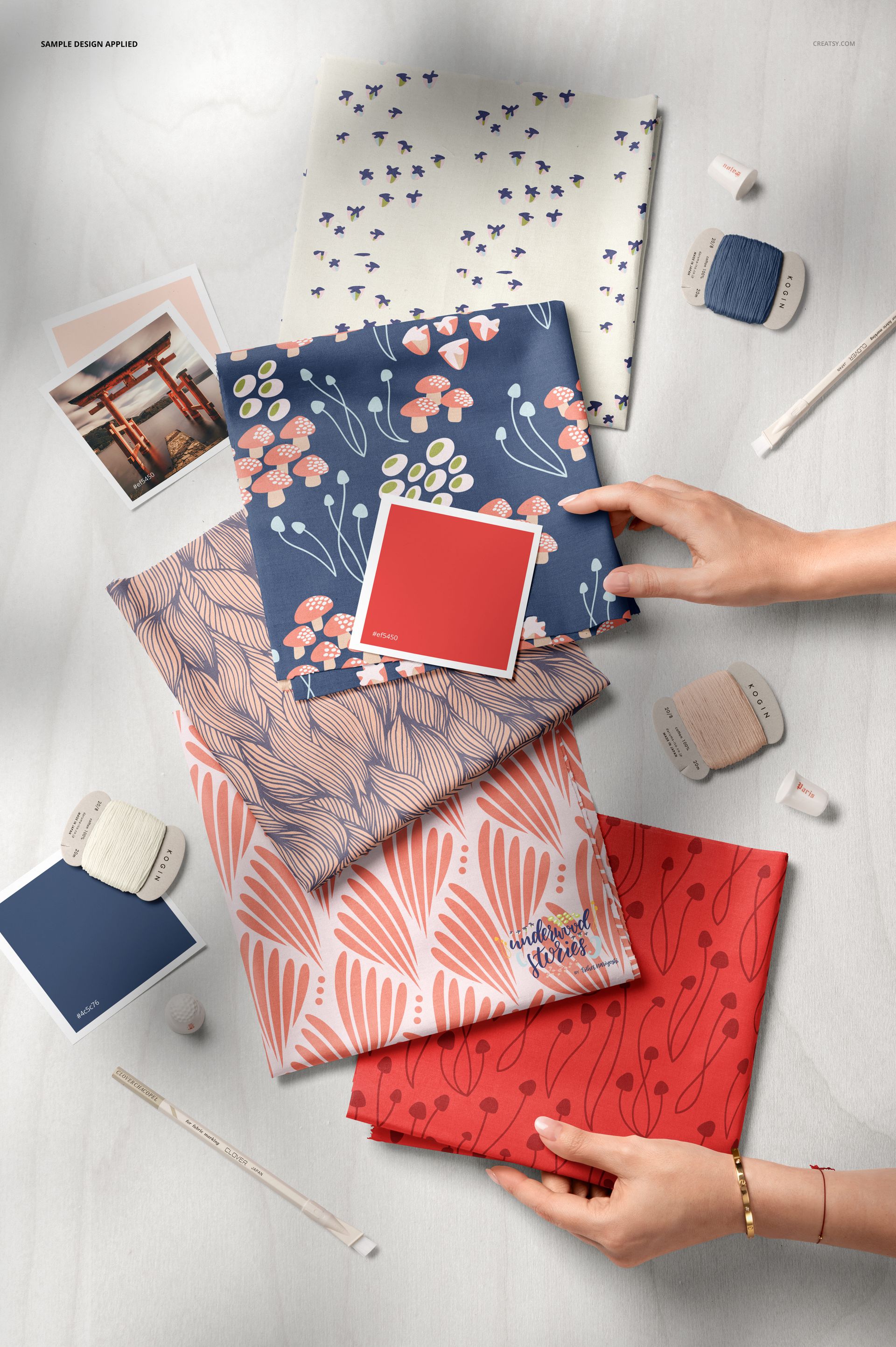 PSD mockup displaying five patterned fabric squares, color sample cards, thread spools, and hands arranging items on a pale background.